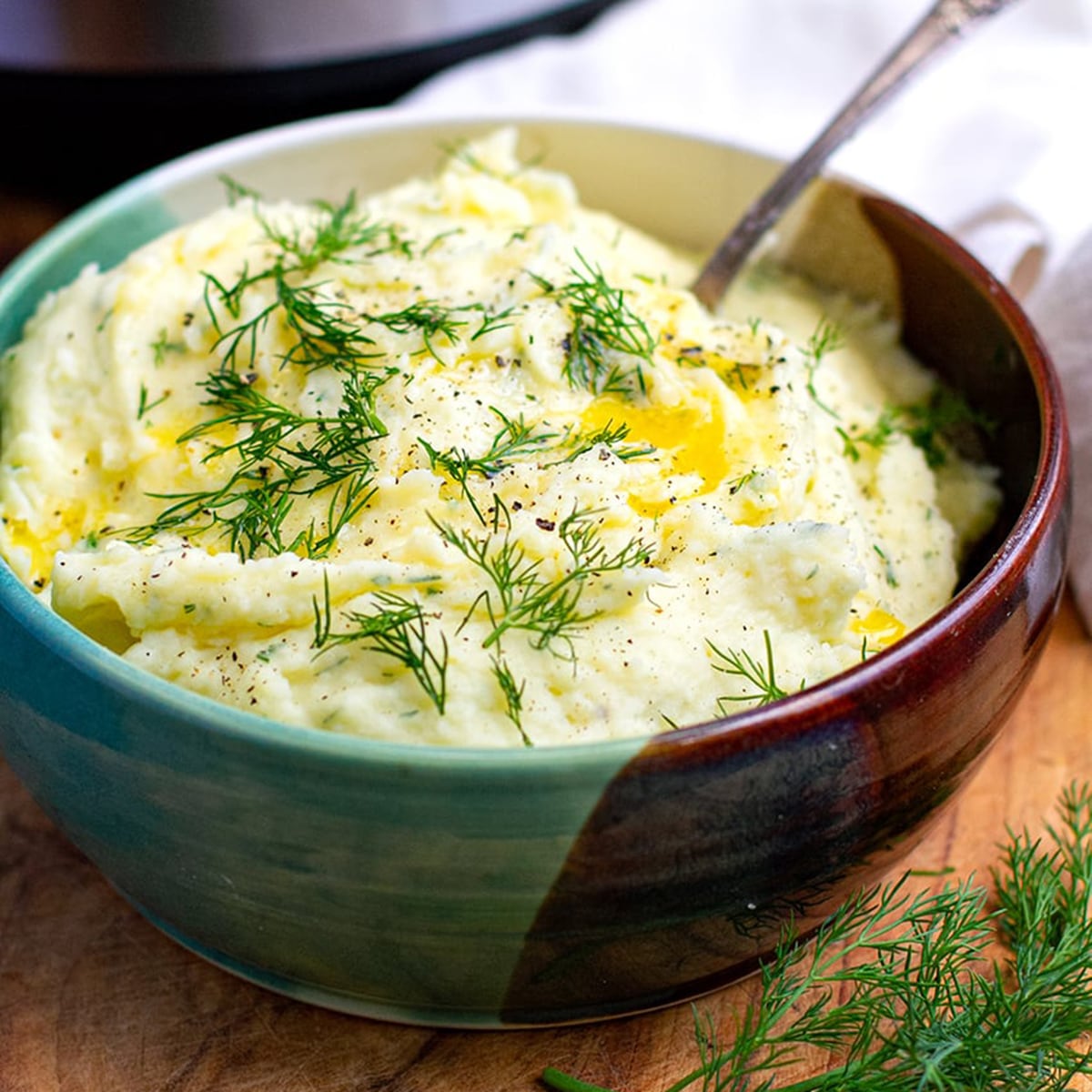 Creamy cream cheese Instant Pot mashed potatoes drizzled with melted butter and topped with fresh dill herbs in a ceramic bowl for Thanksgiving side dishes for a crowd.