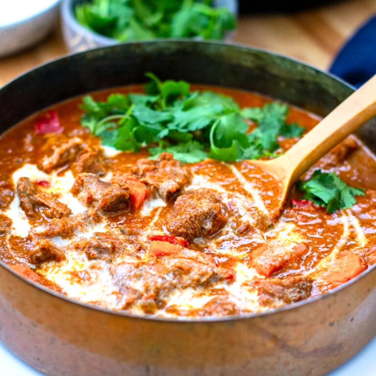 A bowl of rich lamb curry with tomatoes and coconut milk, garnished with fresh cilantro, on a wooden table.