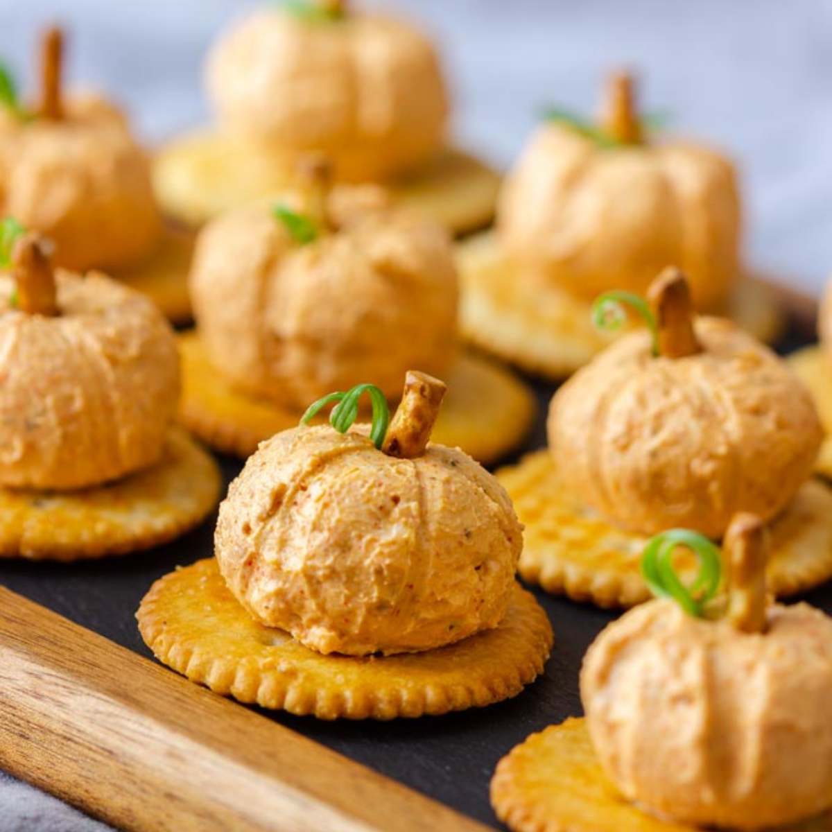  A wooden board filled with miniature pumpkin-shaped cheese balls, each with a pretzel stem, served on crackers.