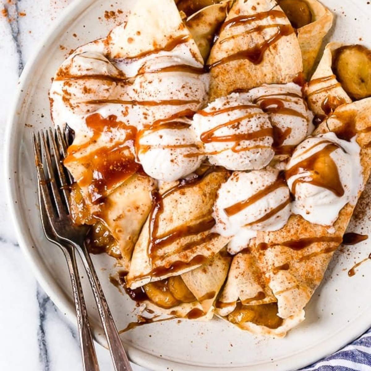 A plate of folded crepes filled with caramelized bananas, topped with vanilla ice cream, caramel sauce, and cinnamon.