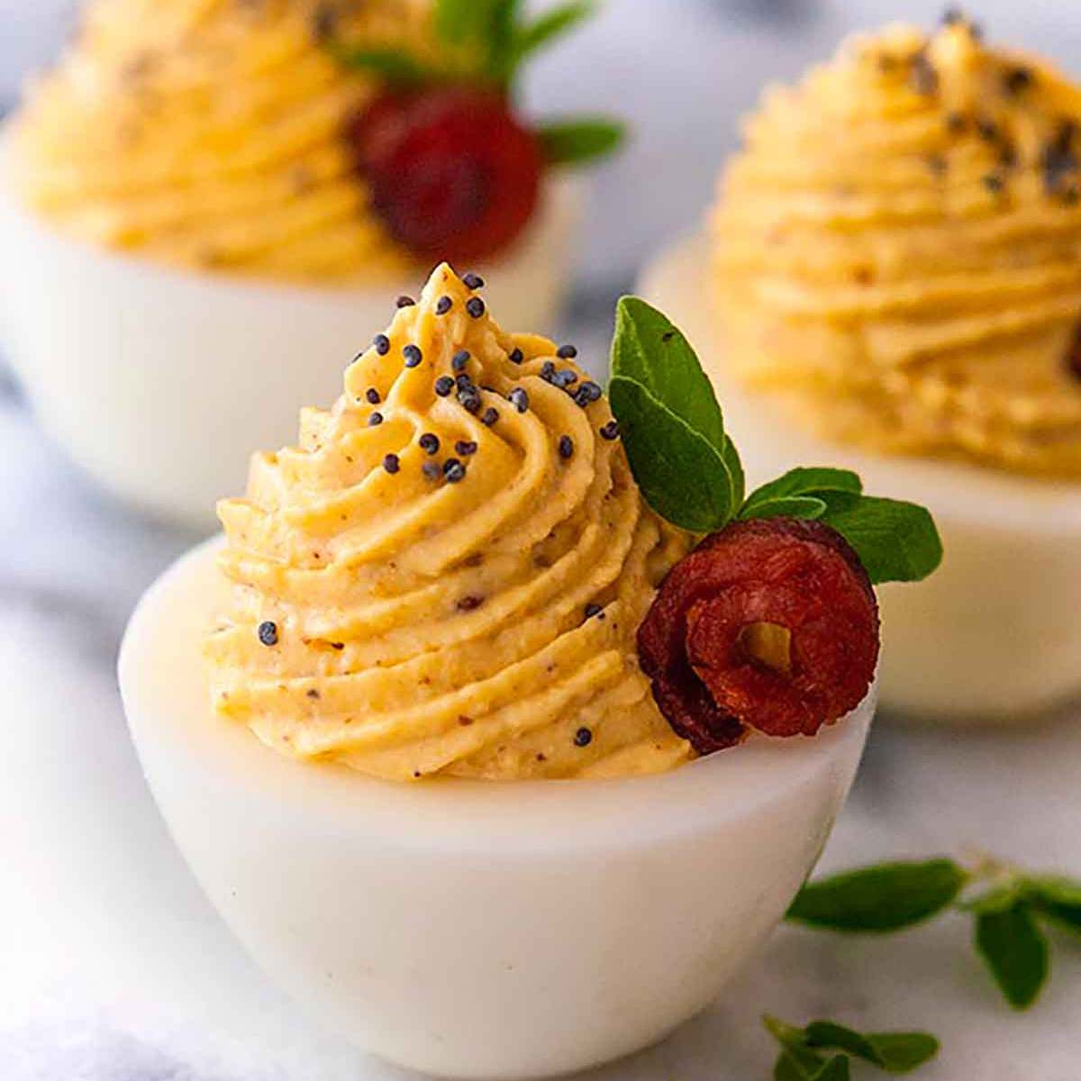 Three deviled eggs with a creamy orange filling, topped with poppy seeds and garnished with fresh thyme and a delicate bacon roses.