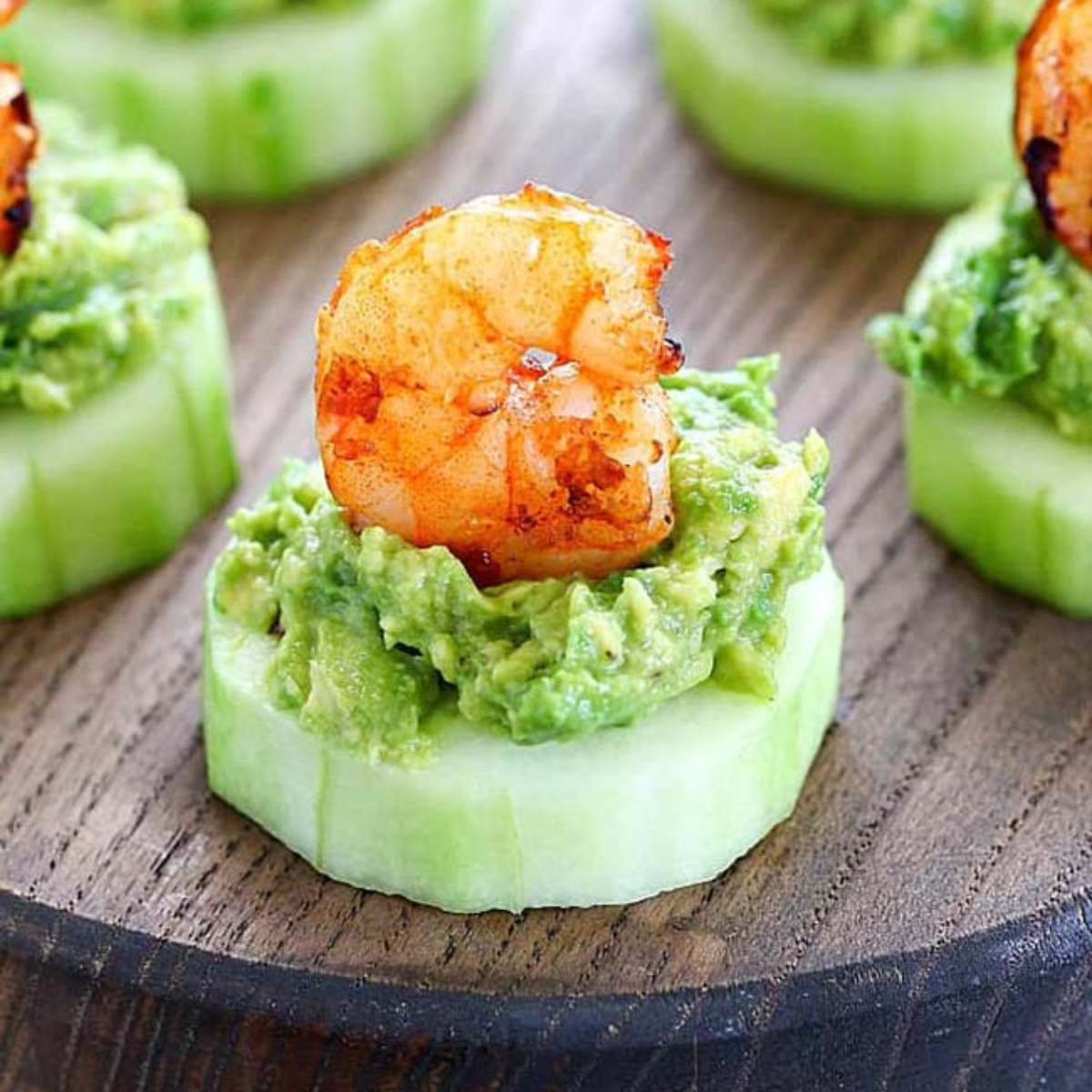  A wooden board with several cucumber slices topped with mashed avocado and grilled shrimp. cold appetizer recipes