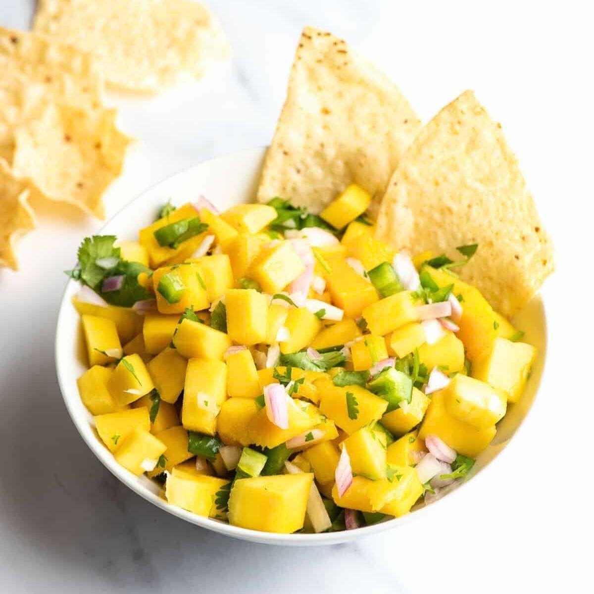  A white bowl filled with fresh mango salsa, made with diced mango, red onion, cilantro, jalapeño peppers, and lime juice. Served with tortilla chips.