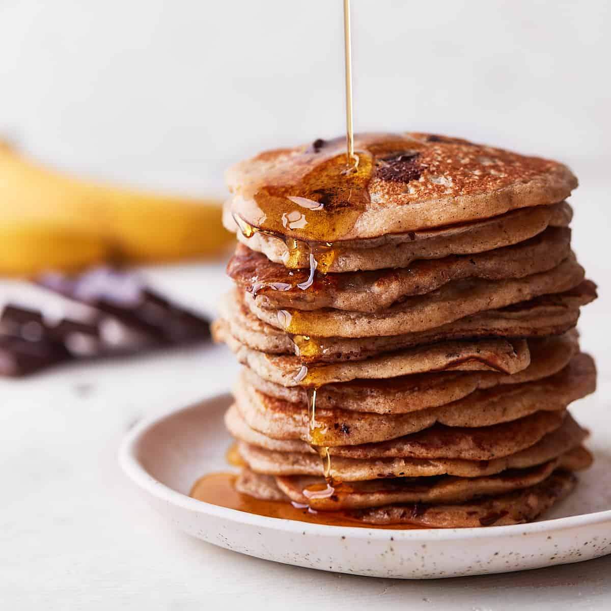 A stack of banana chocolate chip pancakes drizzled with maple syrup, with a banana and chocolate bar in the background.