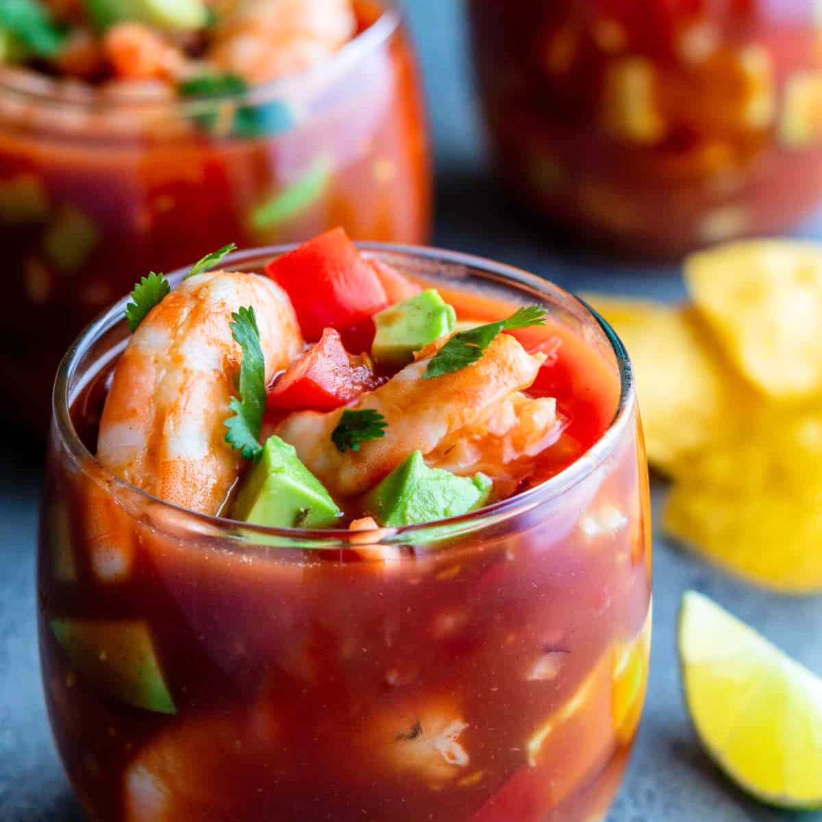 A glass full of shrimp cocktail, filled with shrimp, tomatoes, avocado, and cilantro with tortilla chips and a lime wedge in the background.