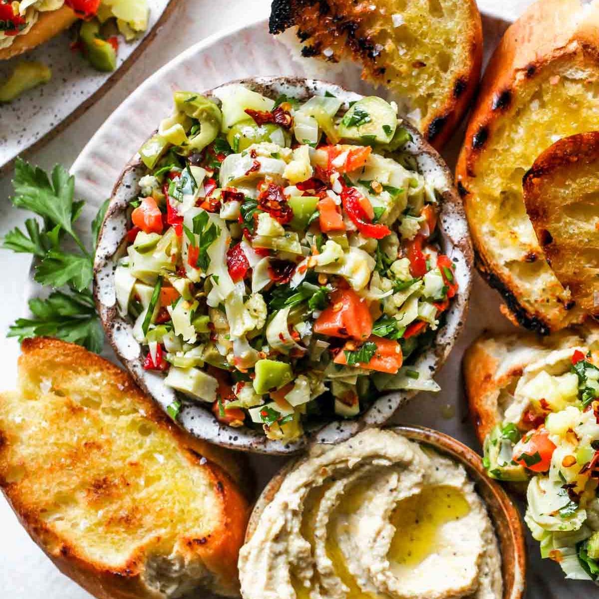  A bowl of marinated artichoke hearts, red bell peppers, and olives, served with grilled bread and a small bowl of hummus.