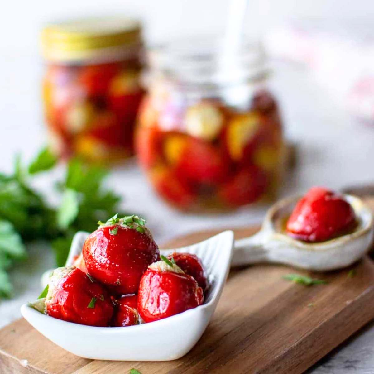 Stuffed cherry peppers, with additional peppers visible in jars and parsley in the background, served in a white bowl.