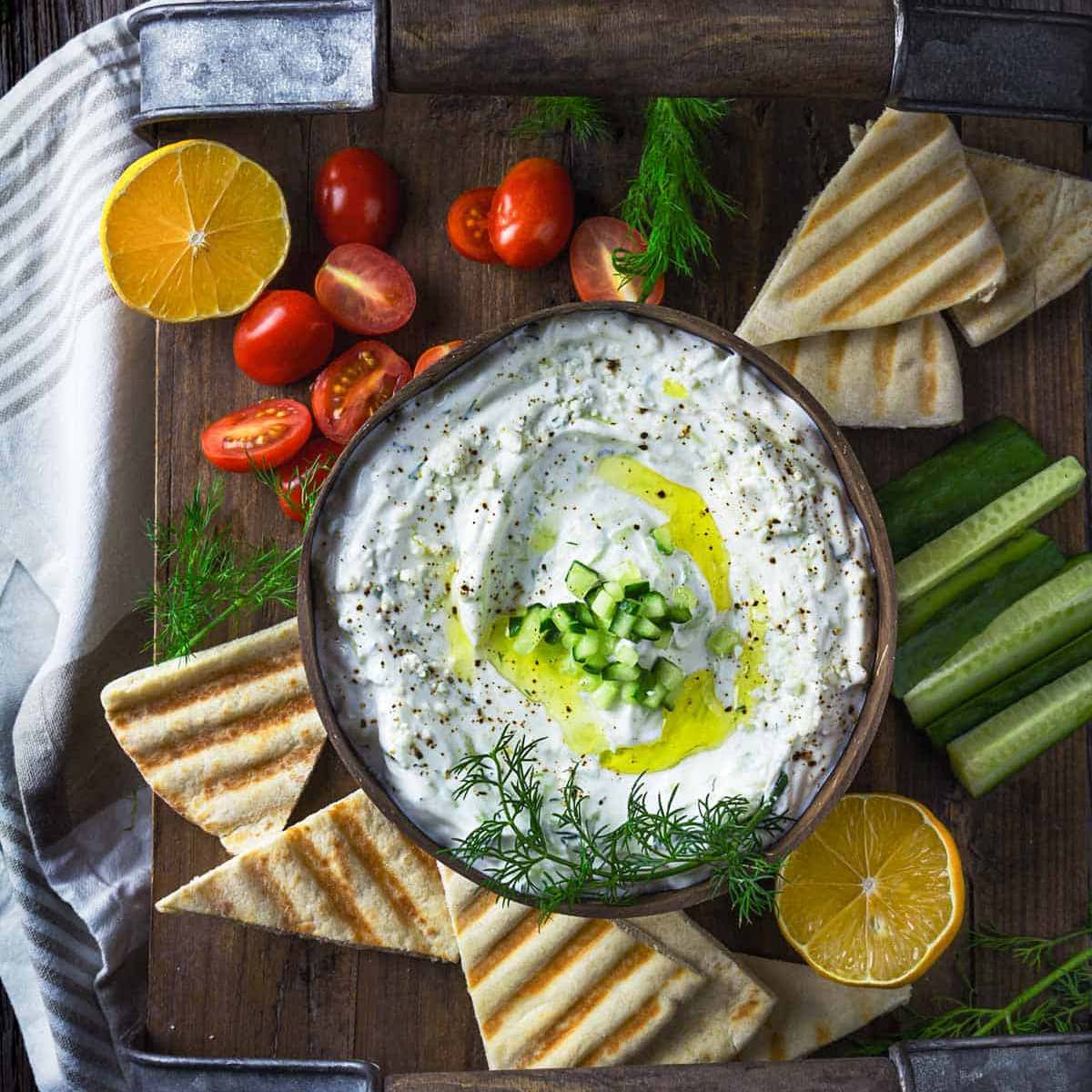  A wooden tray filled with tzatziki sauce, surrounded by pita bread, cucumber slices, cherry tomatoes, lemon wedges.