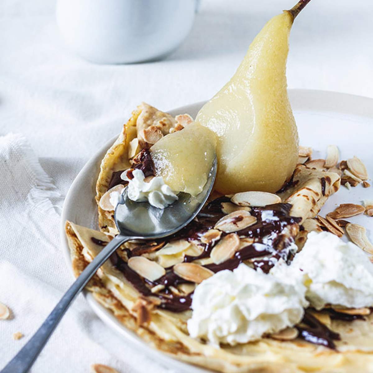 Crepes topped with a poached pear, whipped cream, chocolate sauce, and sliced almonds served on a white plate.