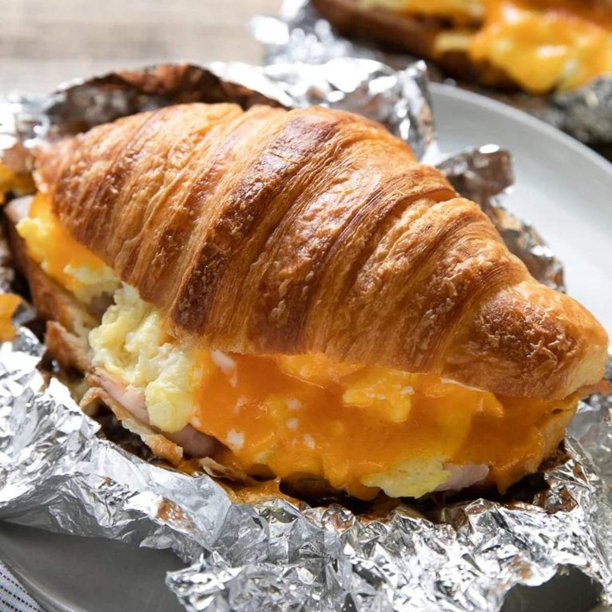 A grilled croissant sandwich filled with scrambled eggs, ham, and melted cheese, wrapped in foil.