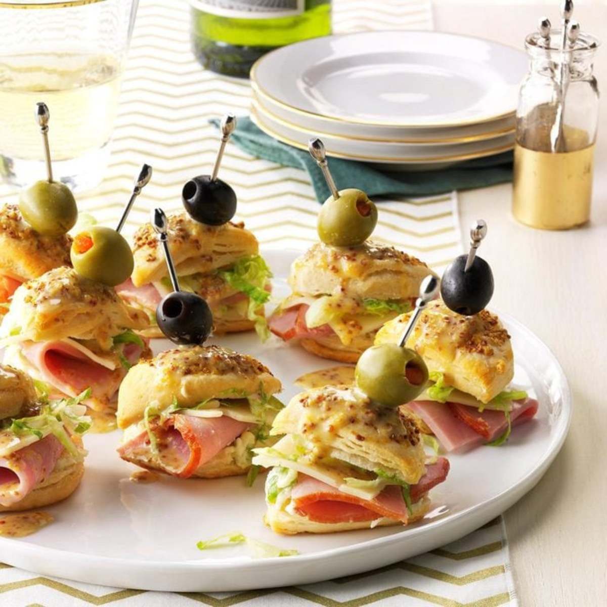  A plate of ham and cheese biscuit stacks, topped with mustard and a green olive, served on a white platter.