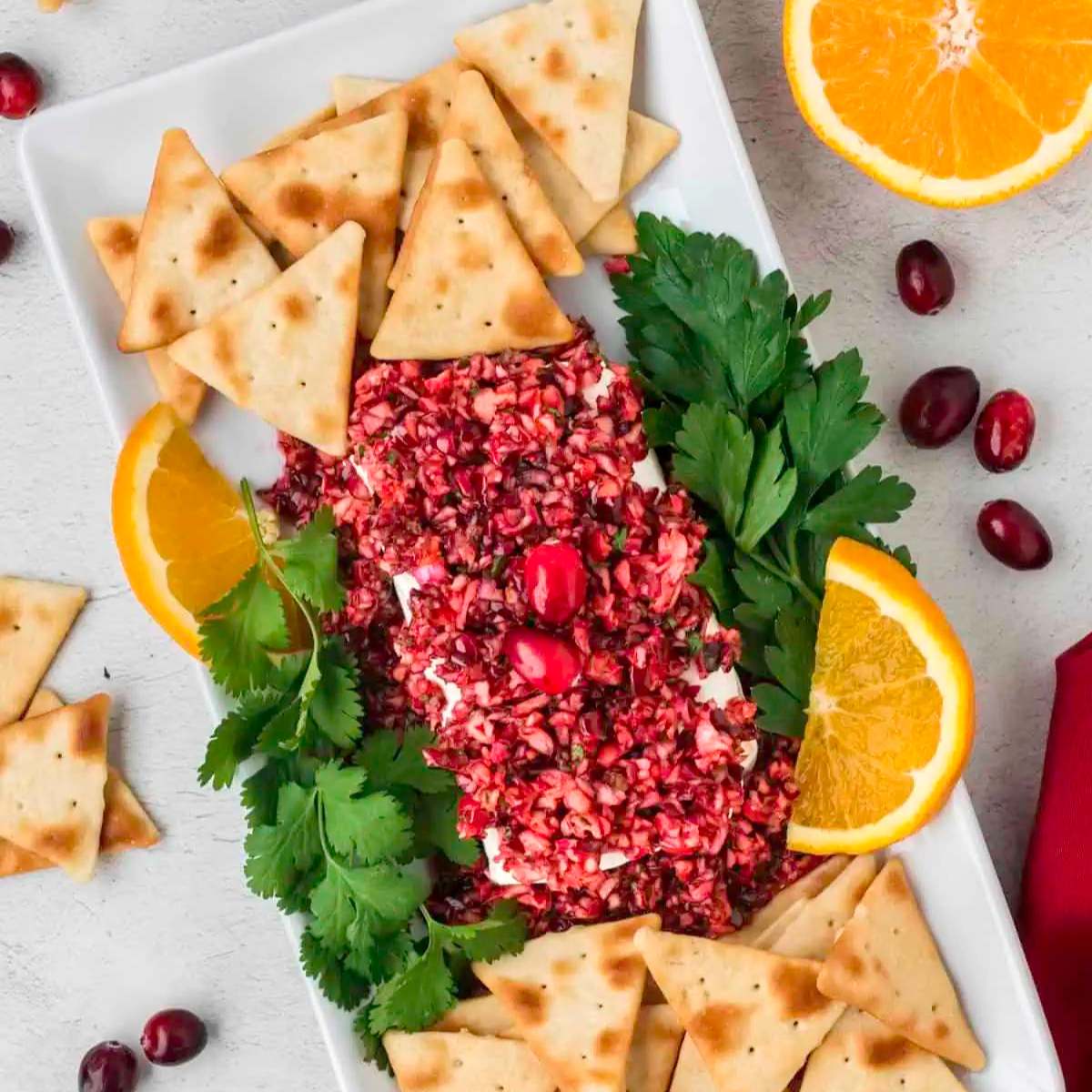 A white platter filled with a topped with a cranberry relish. Surrounded by crackers, orange slices, and fresh parsley.