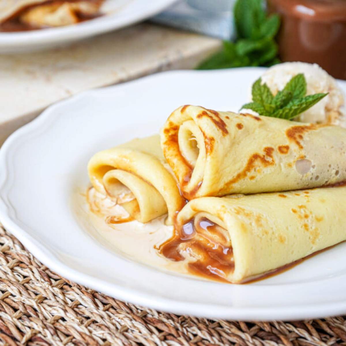 A close-up of three delicate, golden-brown crepes filled with rich Dulce de Leche, neatly rolled and arranged on a white plate.