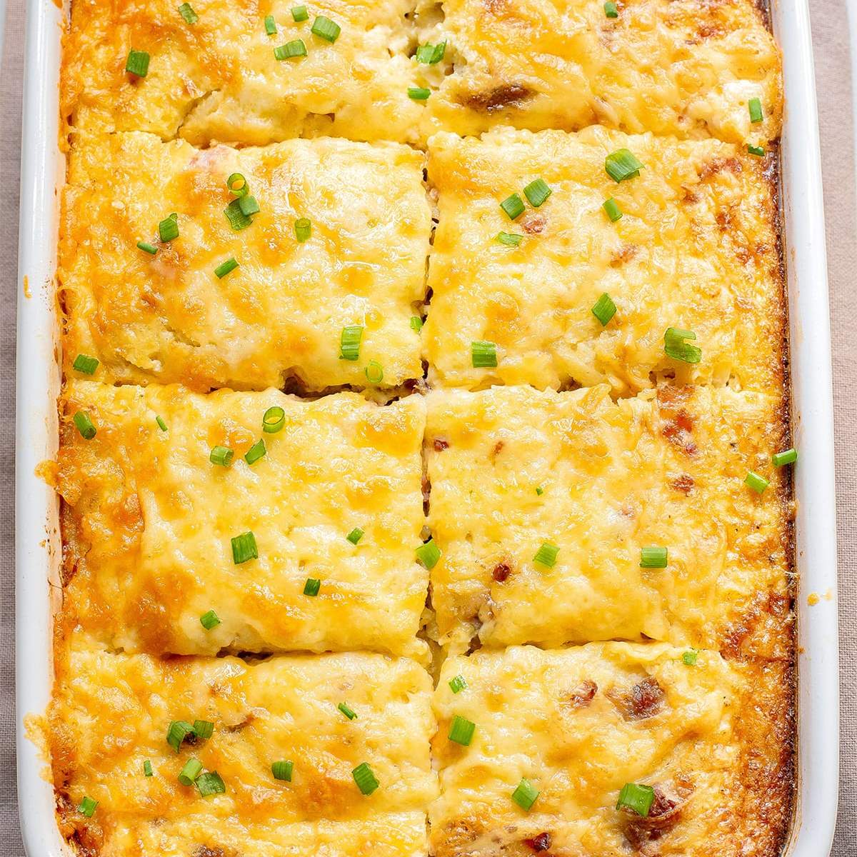 Cheesy potato casserole in a white baking dish, cut into squares. The top is golden brown and bubbly, sprinkled with chopped green onions. 