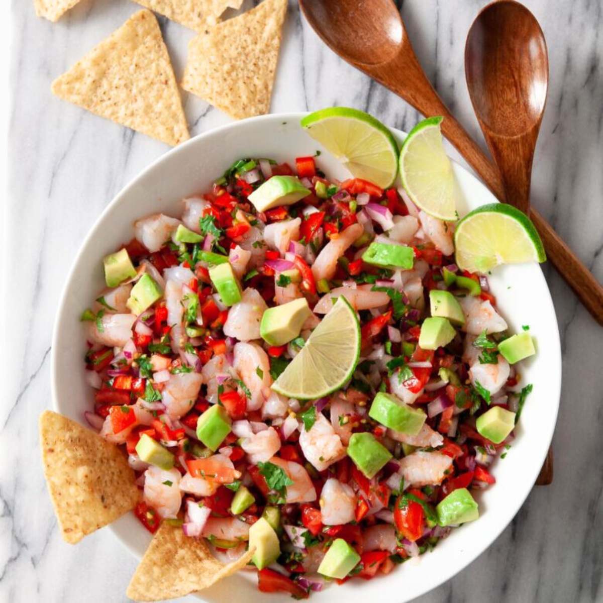 A white bowl filled with shrimp ceviche, made with diced shrimp, tomatoes, red onion, avocado, cilantro, and lime wedges. 