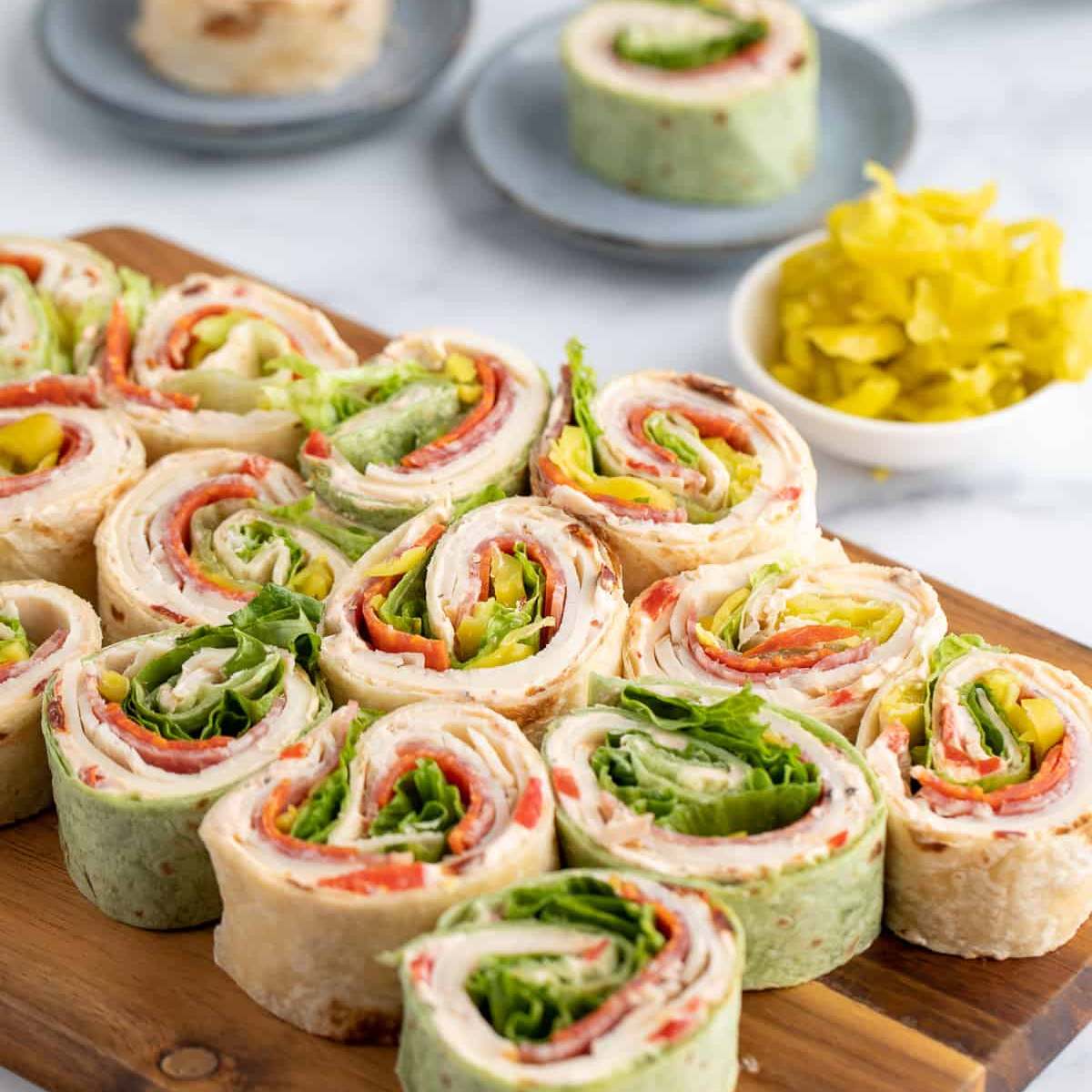 A wooden board filled with colorful pinwheel sandwiches made with tortillas, deli meat, cheese, lettuce, and pepper.