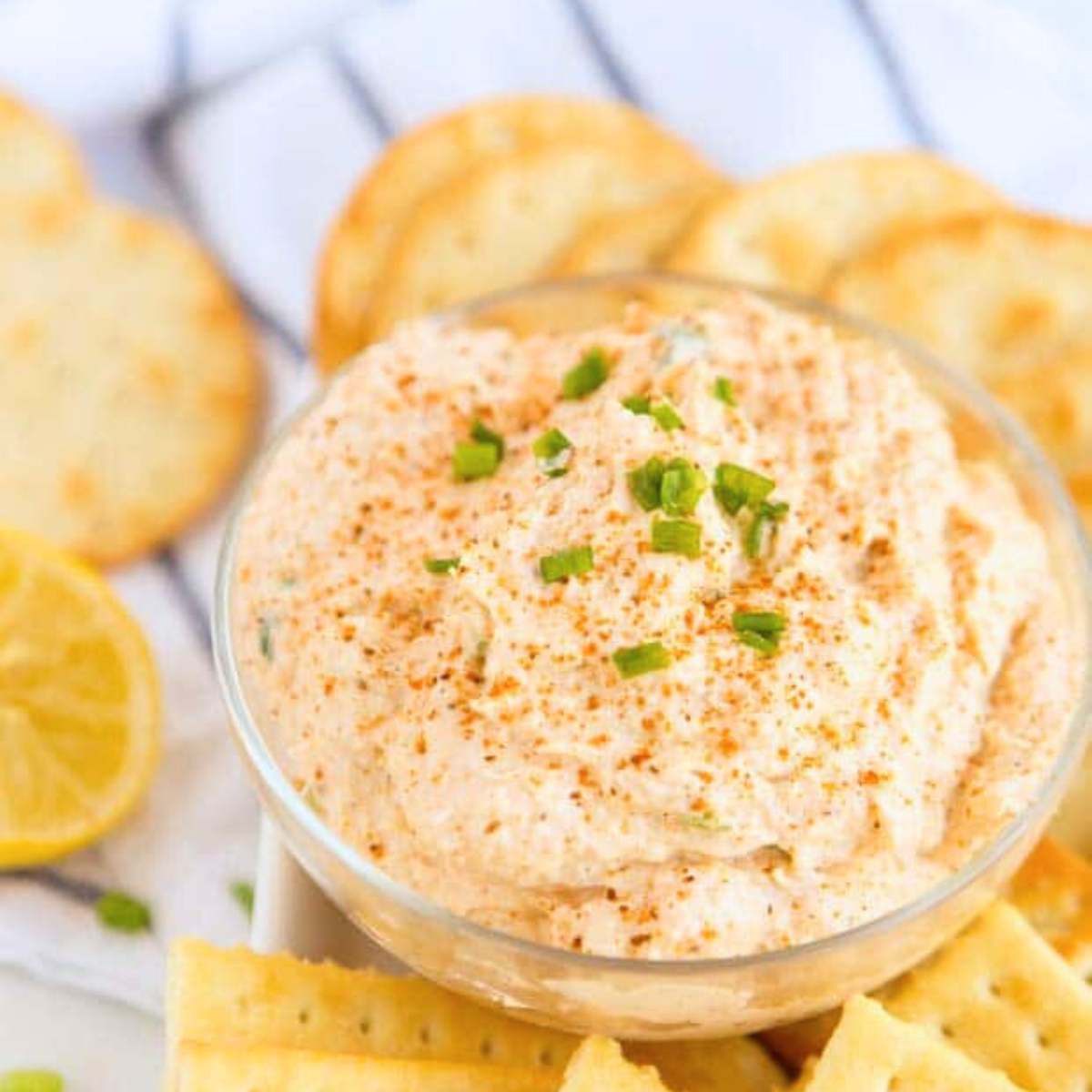 A bowl of creamy crab dip with chopped chives on top, served with crackers and lemon wedges.