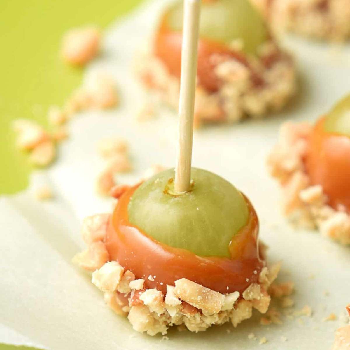 An apple grape on a stick, coated in caramel and chopped peanuts with other caramel apple grapes in the background.