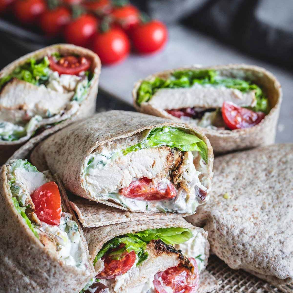 Several Greek chicken wraps cut in half, showing grilled chicken, lettuce, tomatoes, and a creamy sauce inside whole-wheat tortillas.