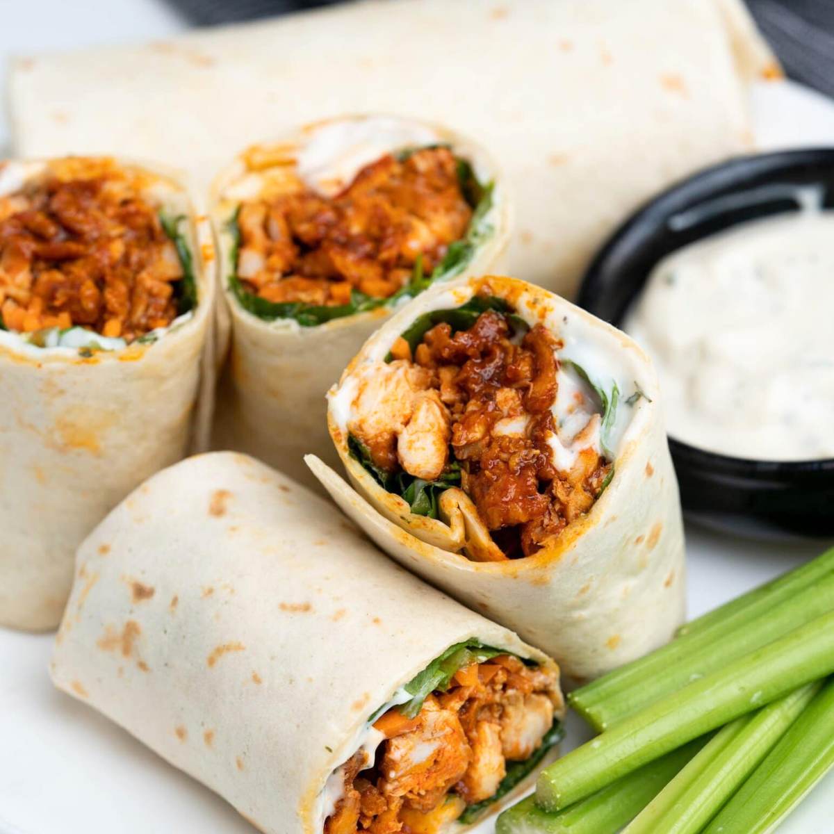  Chicken Wraps cut in half, showing shredded buffalo chicken, lettuce, and a creamy sauce inside tortillas. A small bowl of blue cheese dressing and celery sticks are also on the plate.