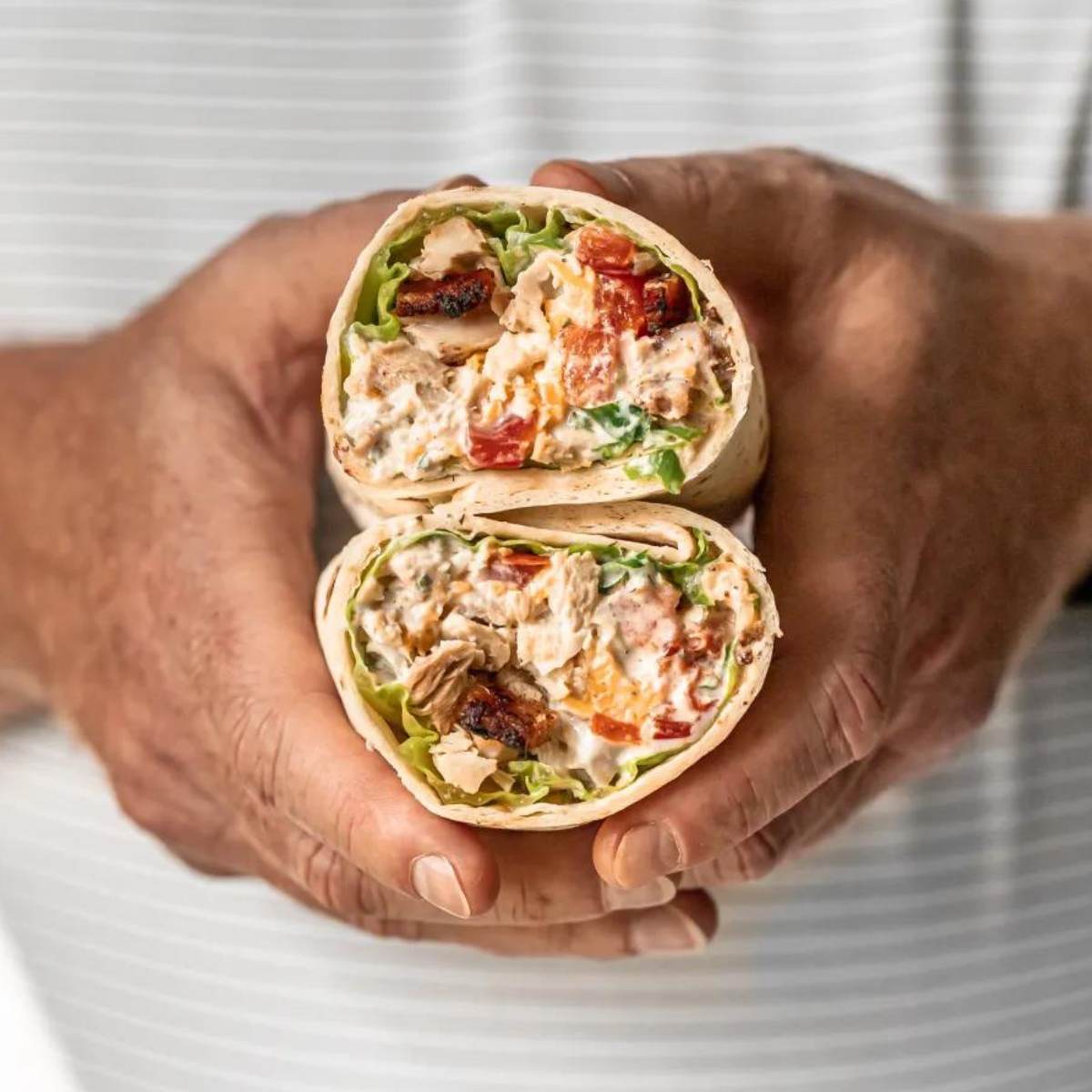  A photo of a person's hands holding a chicken bacon ranch wrap cut in half. The wrap contains grilled chicken, bacon, lettuce, tomato, and a creamy ranch dressing.