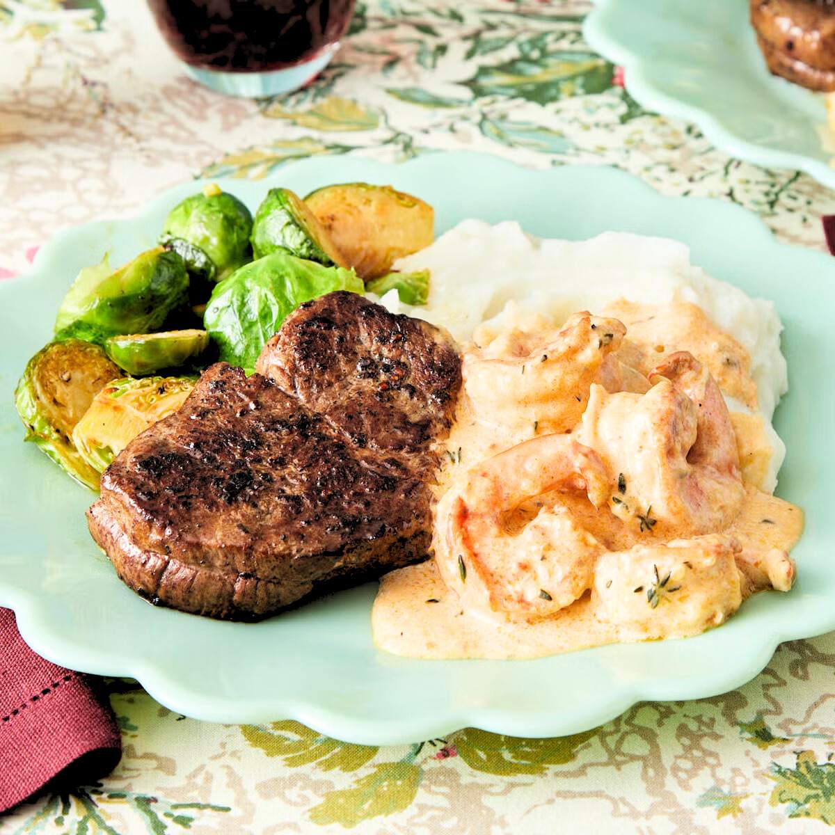  A plate of surf and turf with a seared steak, sautéed shrimp in a creamy sauce, mashed potatoes, and roasted Brussels sprouts.