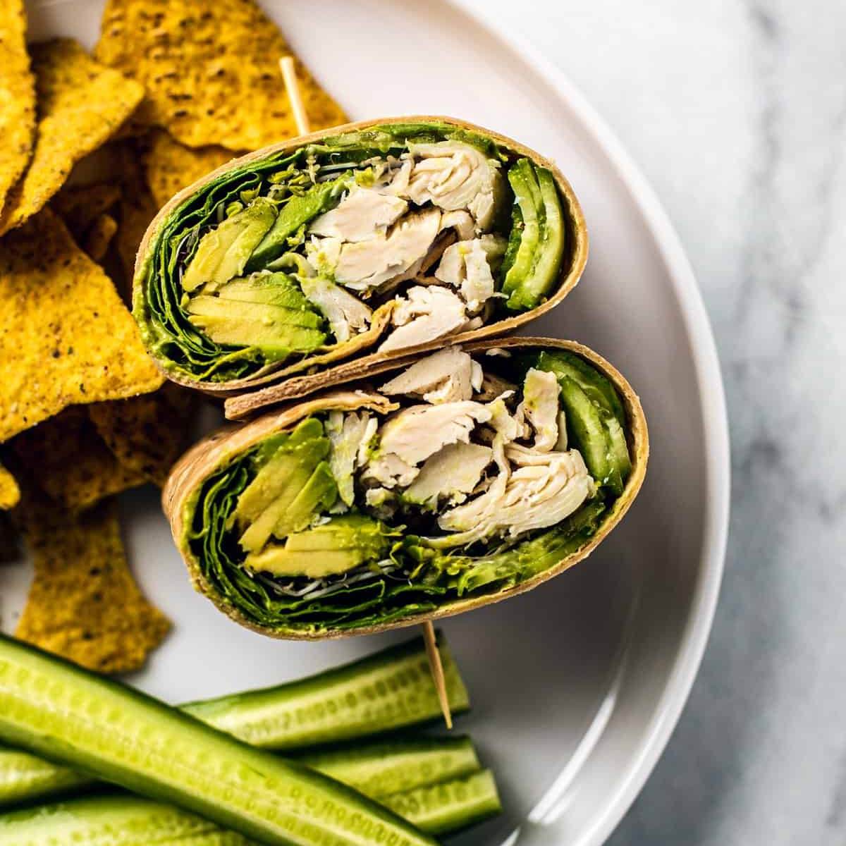 A photo of a Chicken Wrap cut in half, showing shredded chicken, avocado, lettuce, and a creamy dressing inside a whole-wheat tortilla. A side of tortilla chips and cucumber slices is also on the plate.