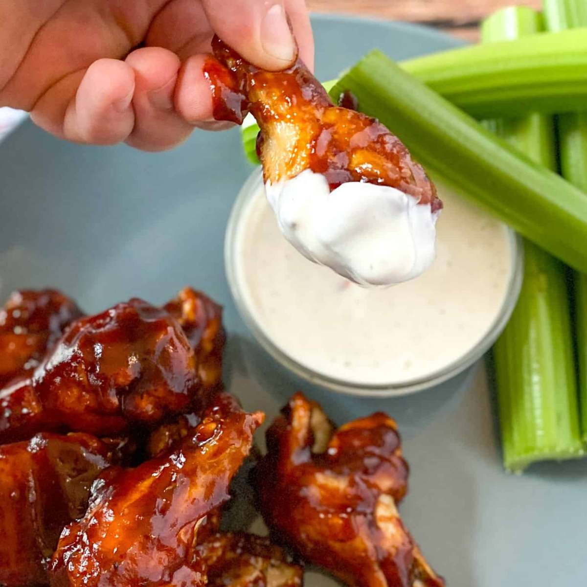 A hand dipping a crispy, saucy chicken wing into a bowl of blue cheese dressing, with celery sticks on the side.