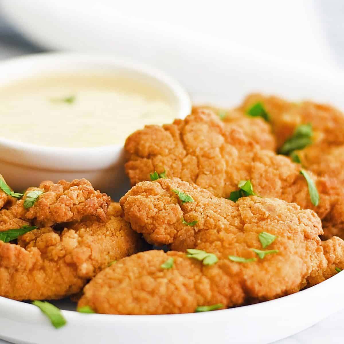 Crispy chicken strips served on a plate with a small bowl of dipping sauce and garnished with fresh parsley.