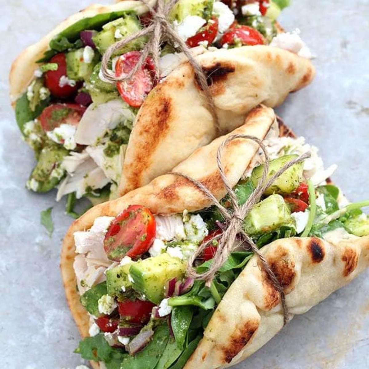 Three Greek Chicken Pitas tied with twine, showing shredded chicken, tomatoes, cucumbers, feta cheese, and a green herb sauce inside the pita bread.