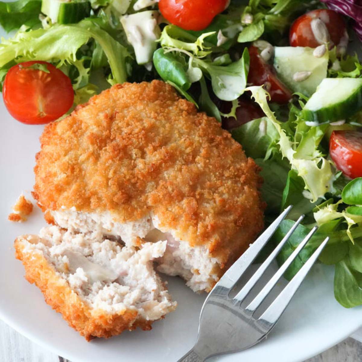 A breaded and fried chicken patty, partially cut open to show the juicy interior, served on a plate with a side salad.