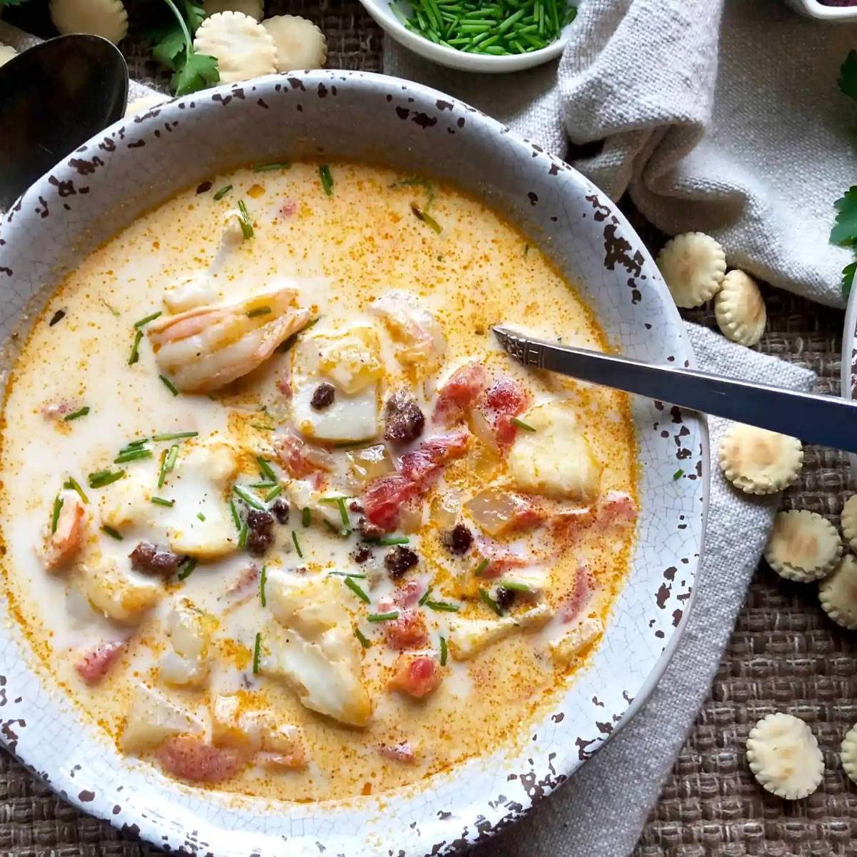 Creamy seafood soup with white fish, shrimp, potatoes, and vegetables, served in a white bowl with crackers on the side.

