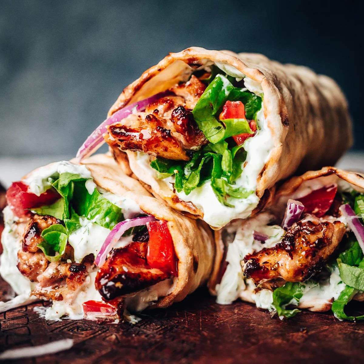 Several chicken wraps stacked on top of each other, showing marinated chicken, lettuce, tomatoes, red onion, and white sauce inside pita bread.