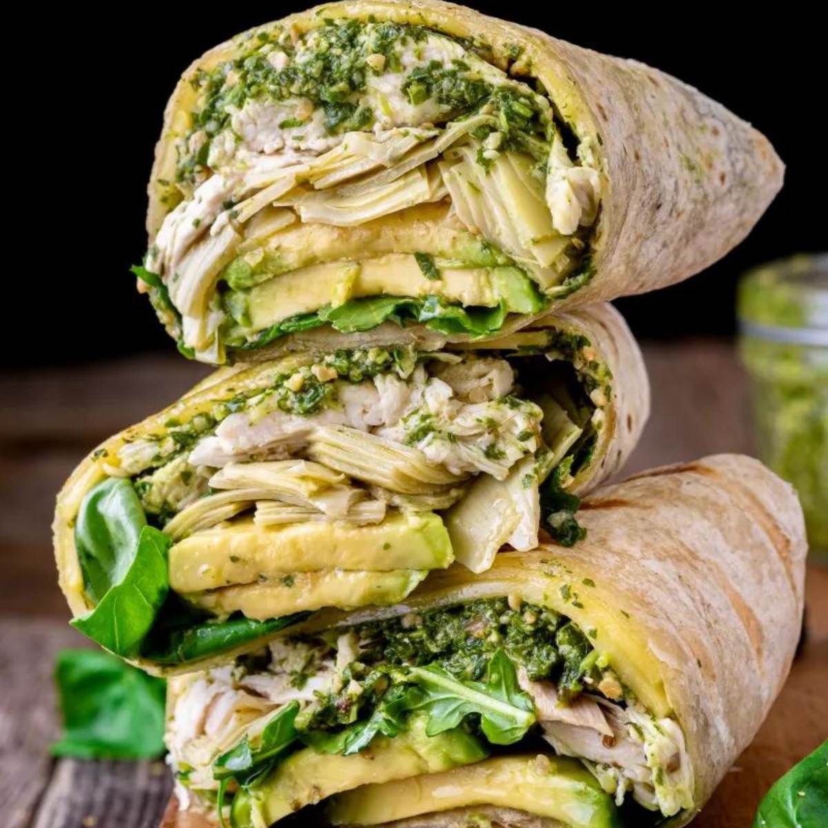 A stack of three Chicken Wraps cut in half, showing grilled chicken, avocado, marinated artichokes, and pesto sauce inside a whole-wheat tortilla.