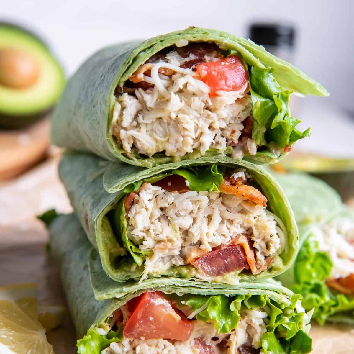  A stack of three chicken wraps cut in half, showing shredded chicken, bacon, avocado, lettuce, and tomato inside a spinach tortilla.