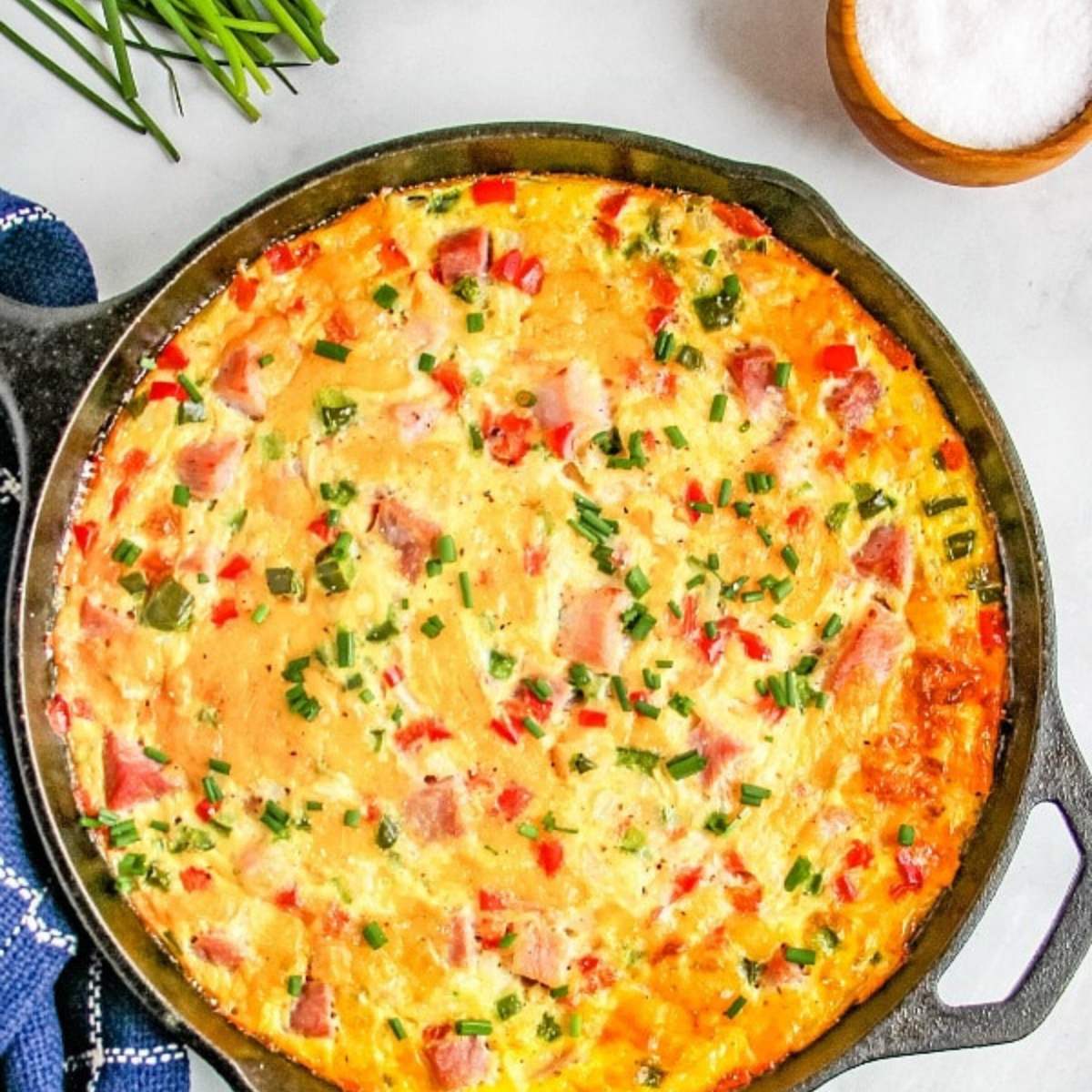 A savory frittata made with eggs, ham, cheese, and vegetables, served in a cast-iron skillet and topped with fresh chives.