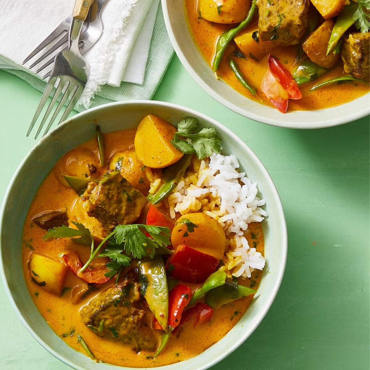 Two bowls of Thai beef and vegetable curry. The curry is a rich, yellow color and contains chunks of beef, potatoes, carrots, and green beans, served with a side of steamed rice and garnished with fresh cilantro.