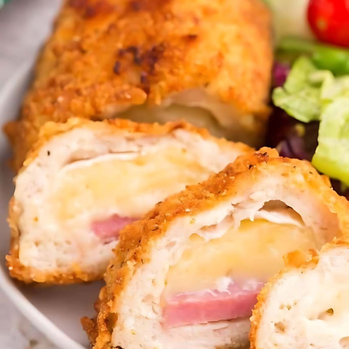 Crispy, golden-brown chicken cordon bleu cut open to reveal layers of ham and cheese. A side salad is visible in the background.