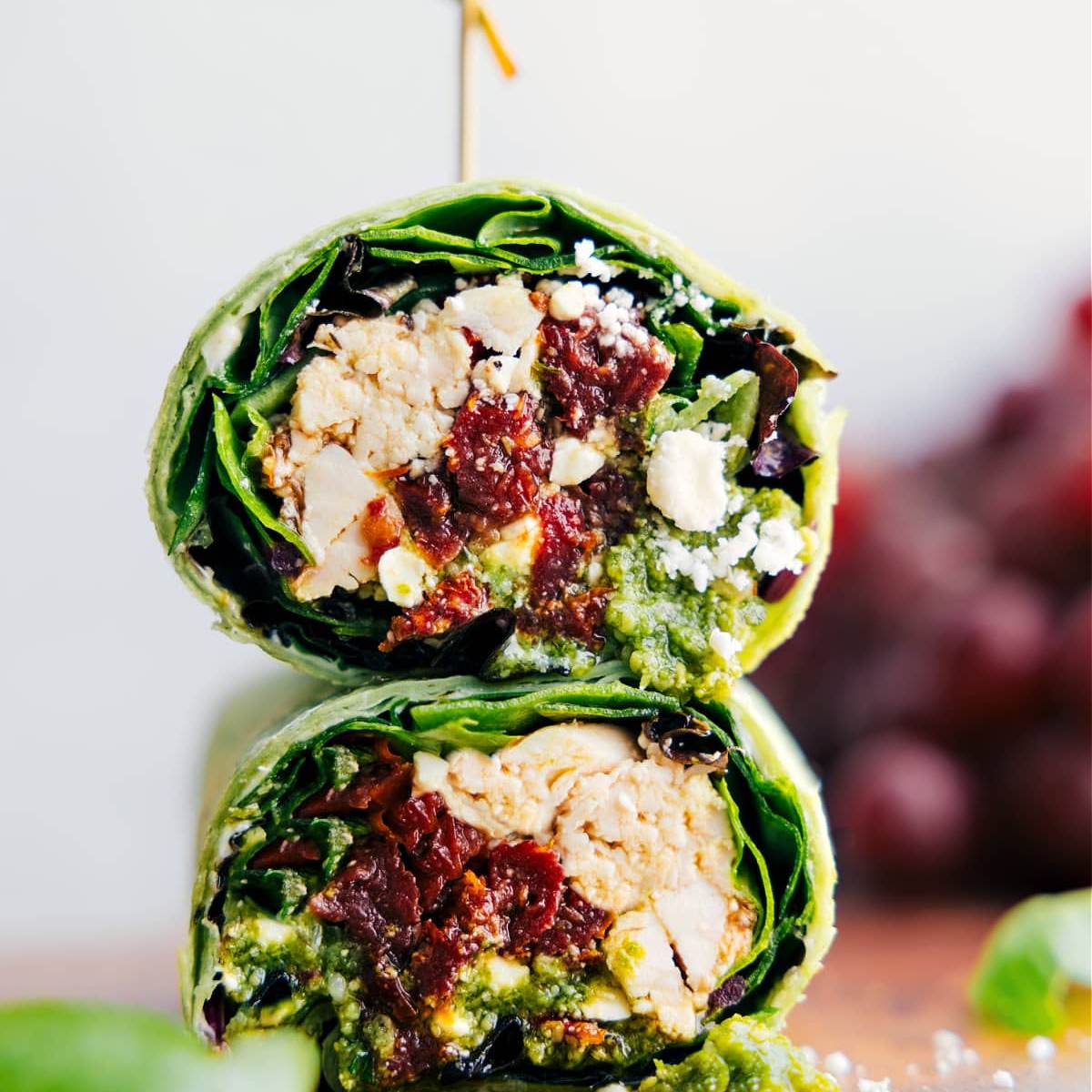 A photo of a chicken wrap cut in half, showing shredded chicken, sun-dried tomatoes, feta cheese, and a pesto sauce inside large spinach leaves.