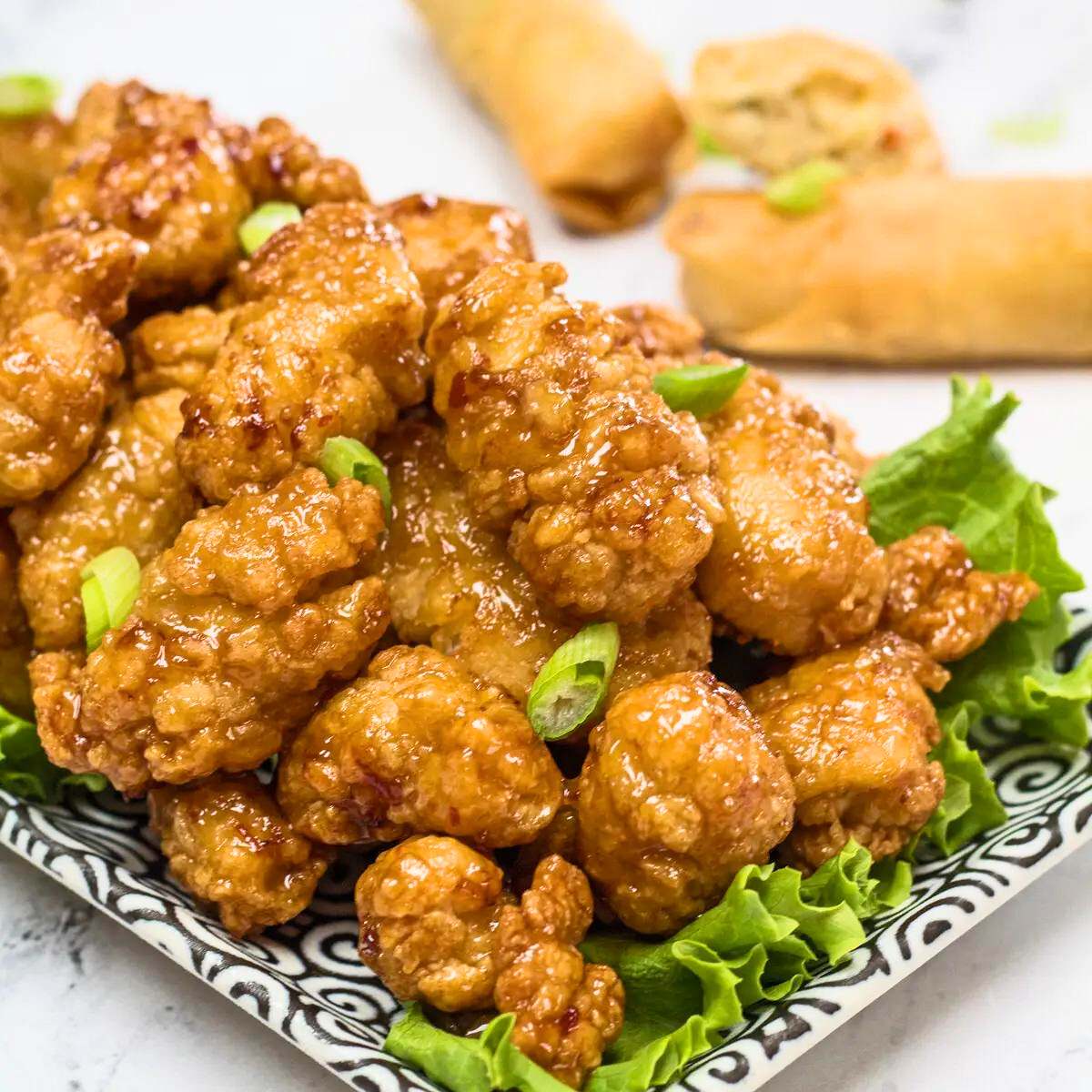 Crispy orange chicken coated in a sweet and tangy sauce, served on a bed of lettuce with spring onions and spring rolls in the background.