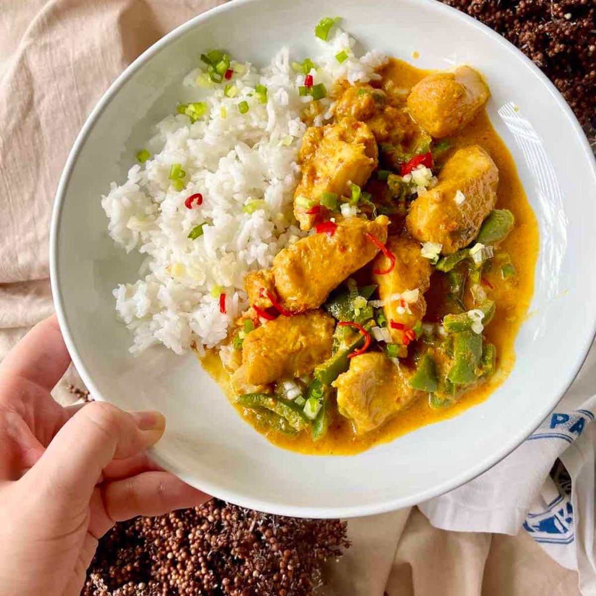  A white bowl filled with a yellow curry, with pieces of fish, green chilies, and red chili flakes. The bowl is held by a hand, and there is a portion of white rice on the side.