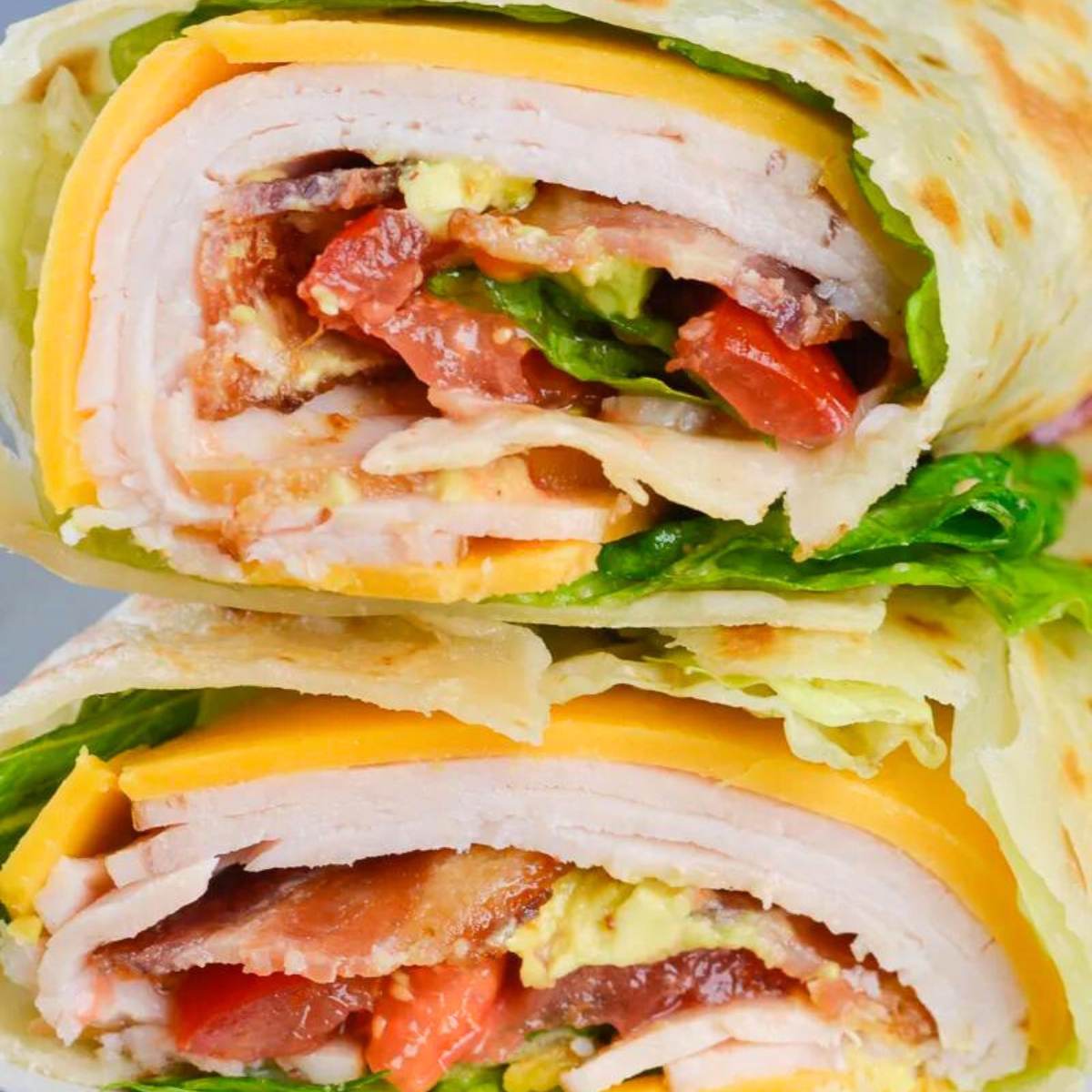 A photo of a Turkey Wrap cut in half, showing turkey, bacon, avocado, lettuce, tomato, and cheese inside a tortilla.