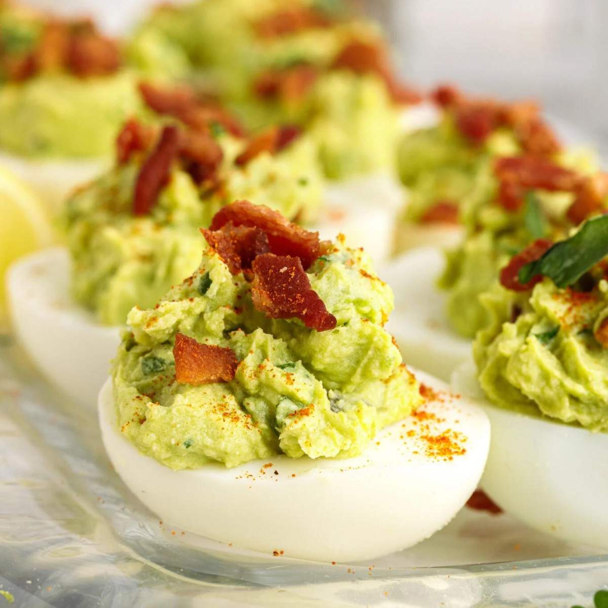 Deviled eggs filled with a creamy avocado mixture, topped with crumbled bacon and paprika.