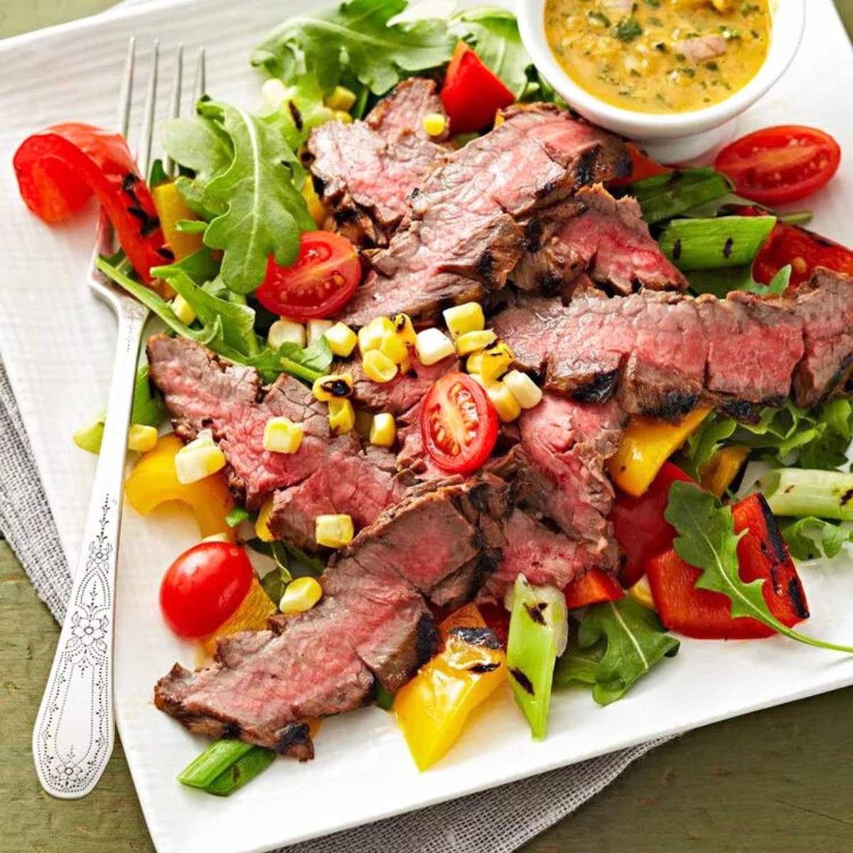 Grilled flank steak salad with mixed greens, grilled bell peppers, cherry tomatoes, corn kernels, and a side of creamy dressing