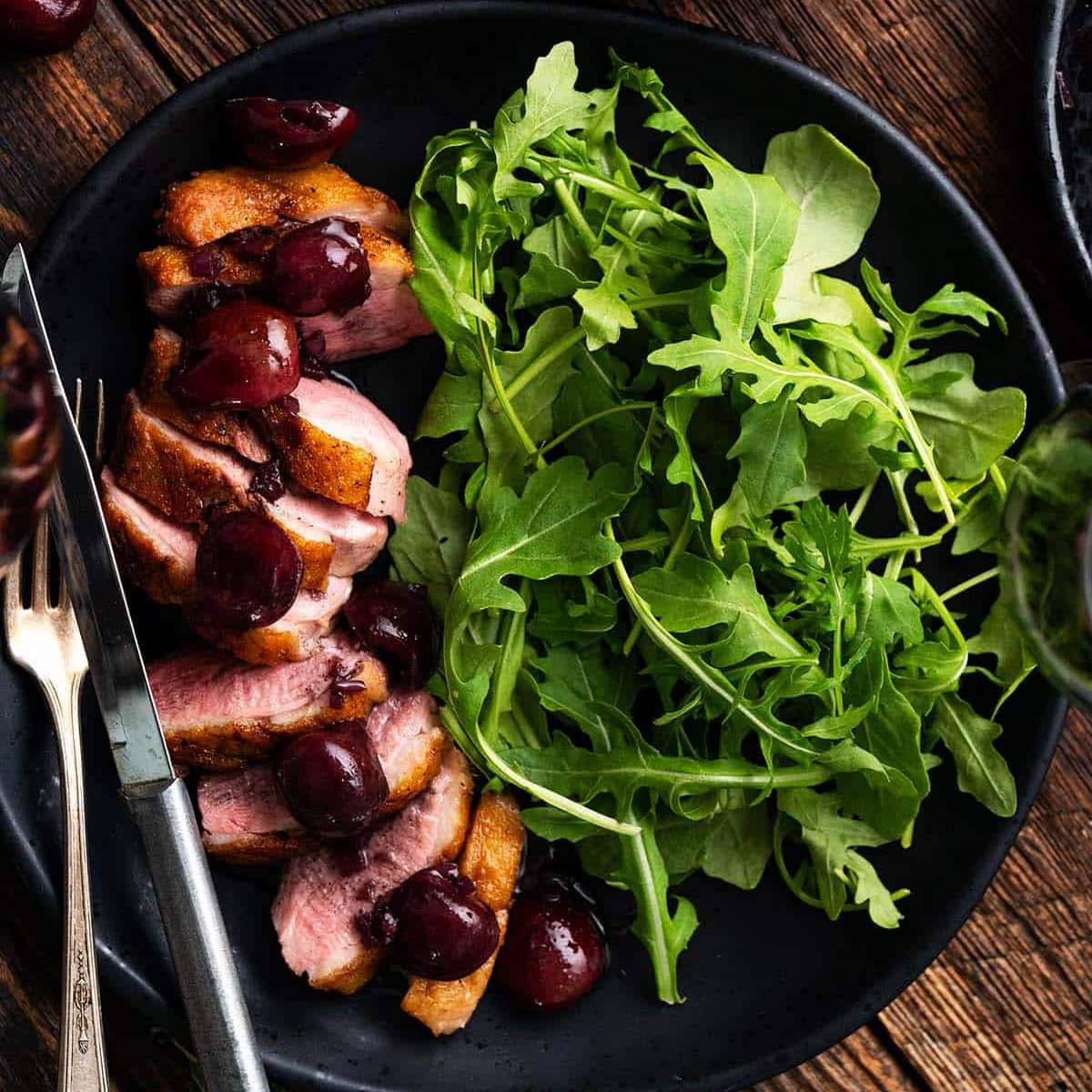 Sliced pan-seared duck breast served with a cherry sauce and a bed of arugula salad, served on a black plate.