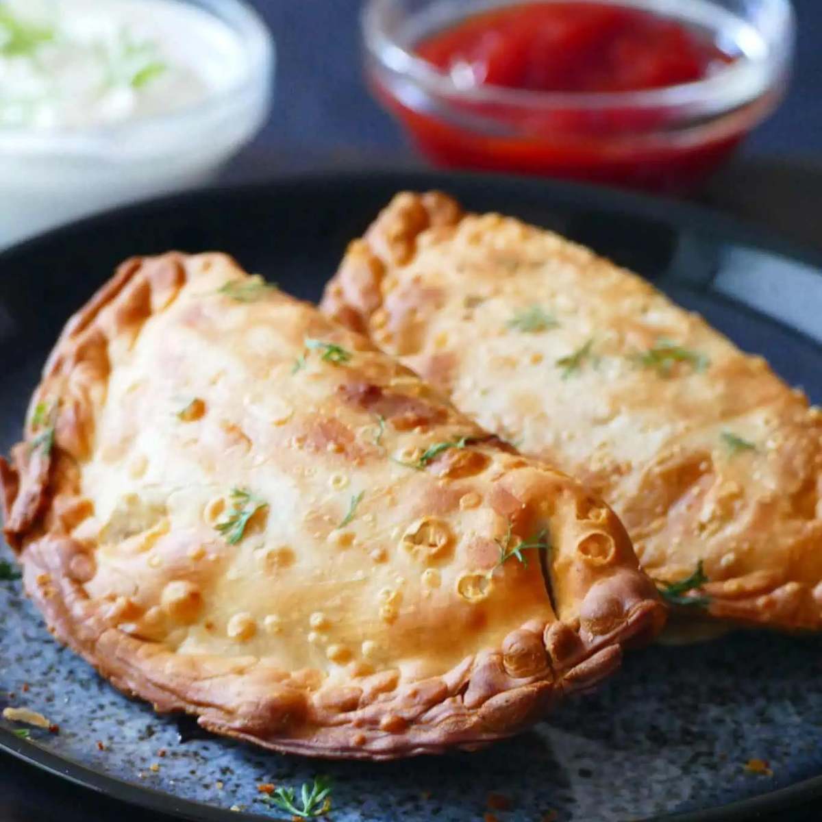 Two golden brown empanadas served on a plate with dipping sauces in the background.