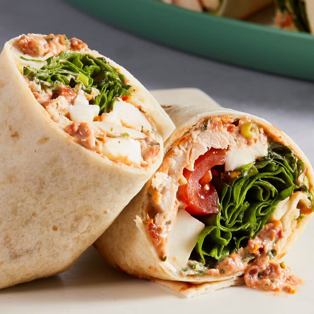 A photo of a Chicken Wrap cut in half, showing shredded chicken, tomato, lettuce, feta cheese, and a creamy spread inside a whole-wheat tortilla.