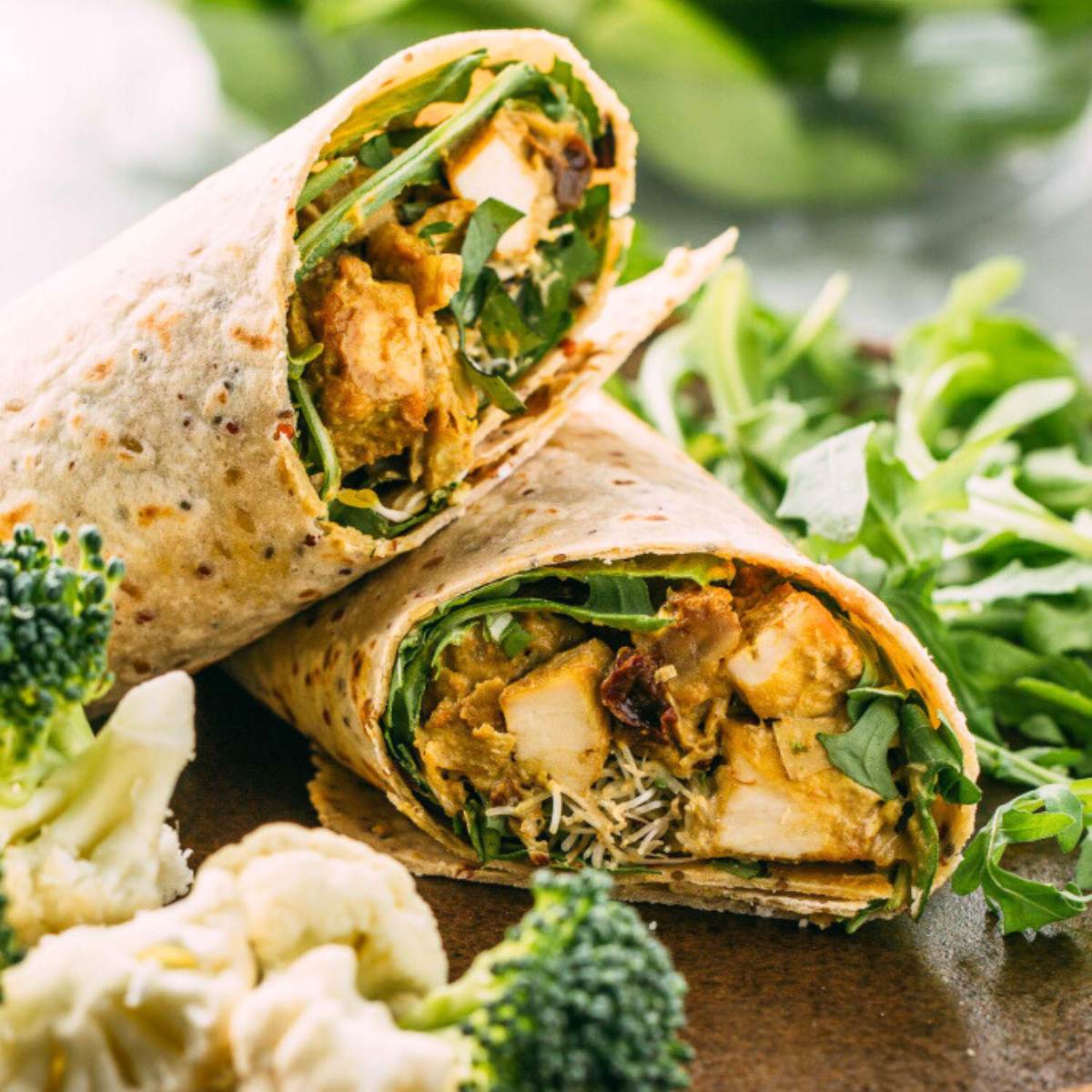 A photo of a Chicken Wrap cut in half, showing shredded chicken, broccoli, spinach, and a creamy coconut curry sauce inside a whole-wheat tortilla. A side of roasted cauliflower is also on the plate.