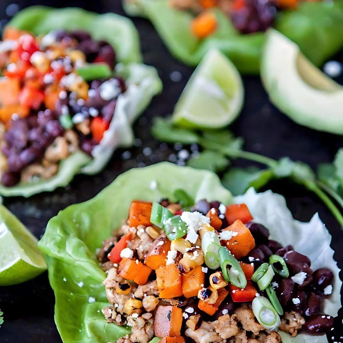 Several lettuce wraps filled with ground chicken, sweet potatoes, black beans, corn, red bell peppers, and shredded cheese. Lime wedges and cilantro are also on the plate.