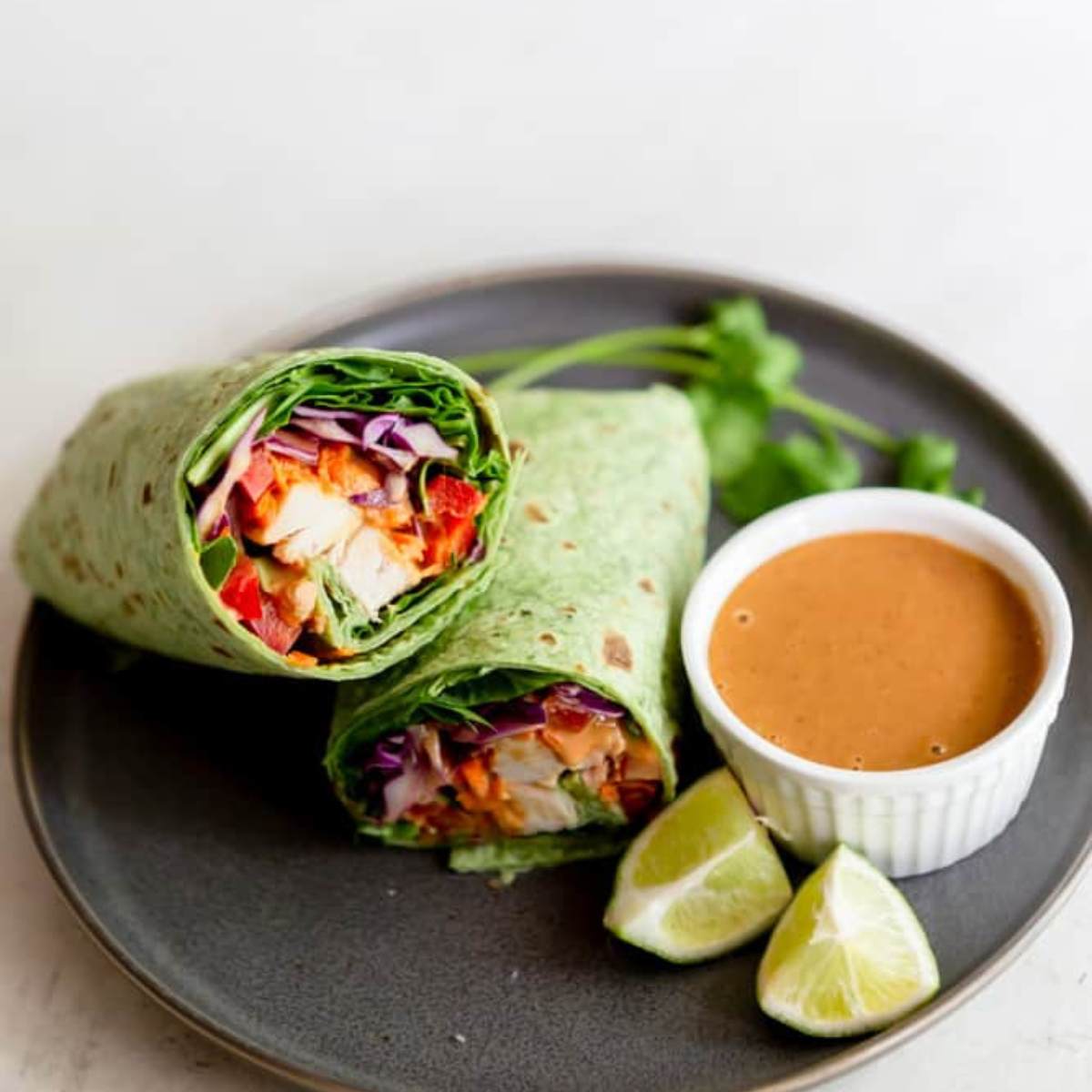 A plate with two chicken wraps cut in half, showing shredded chicken, red cabbage, and a spicy sauce inside large spinach leaves. A small bowl of peanut sauce and lime wedges are also on the plate.